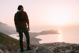 A Caucasian guy stands high on a cliff above a small town and looks at the sun setting behind the islands