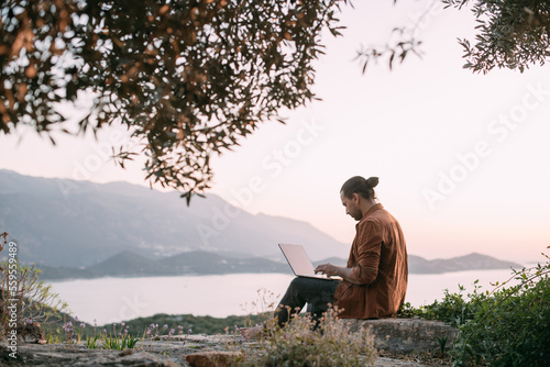 A young Caucasian man remotely works with a laptop in a garden on a mountain overlooking the sea and sunset.