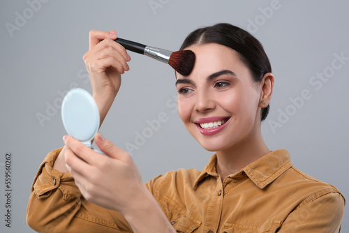 Happy woman with cosmetic pocket mirror applying makeup on light grey background