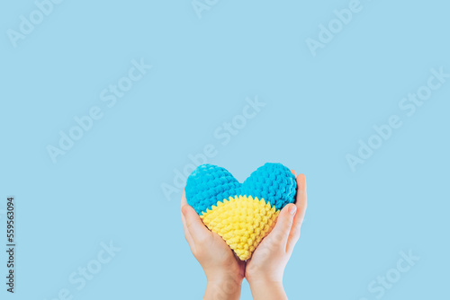 Handmade crochet amigurumi heart in the hands of a child on yellow and blue colors. Flag of Ukraine. Isolated on a blue background. Space for text.