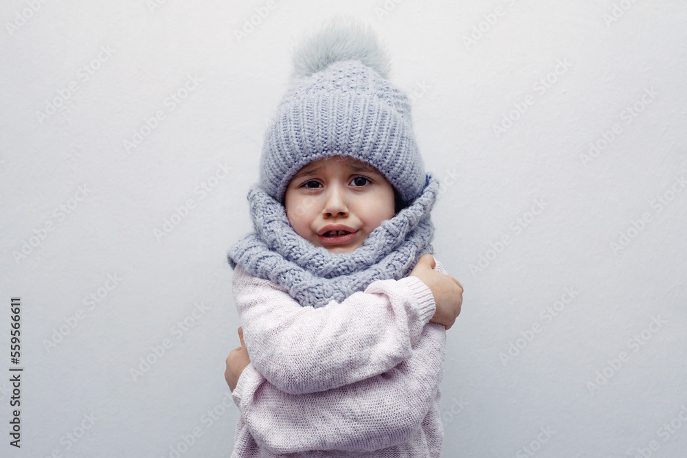 A child in a hat and scarf hugs himself freezing on a white background A child in a hat and scarf hugs himself freezing on a white background