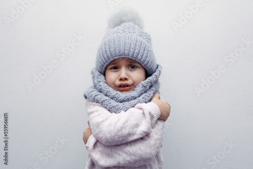 A child in a hat and scarf hugs himself freezing on a white background