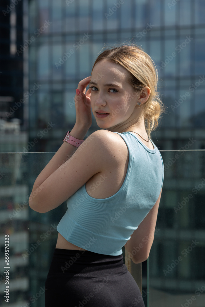 A teenage girl in a blue sports top and black leggings on a terrace among skyscrapers
