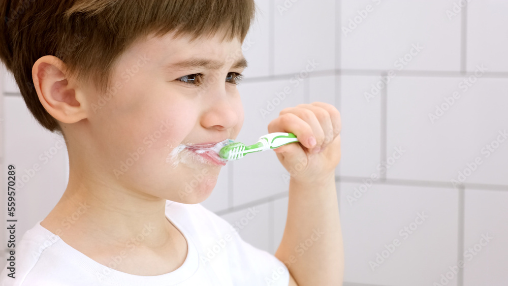 A cute little child brushes teeth. Green toothbrush. Daily morning hygiene procedures in a white bathroom. Portrait of beautiful caucasian 6 year old boy. Teeth brushing concept. Copy space. Close-up A cute little child brushes teeth. Green toothbrush. Daily morning hygiene procedures in a white bathroom. Portrait of beautiful caucasian 6 year old boy. Teeth brushing concept. Copy space. Close-up