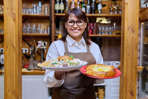 Middle aged female restaurant owner in apron with plates of cooked food