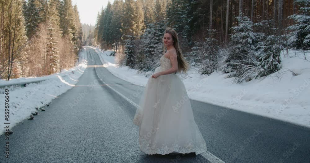 Beautiful Caucasian bride in wedding dress long hair walking running dancing on asphalt road among winter snowy pine forest. Gorgeous young blonde woman alone in dress in winter in Ukraine slow motion Beautiful Caucasian bride in wedding dress long hair walking running dancing on asphalt road among winter snowy pine forest. Gorgeous young blonde woman alone in dress in winter in Ukraine slow motion