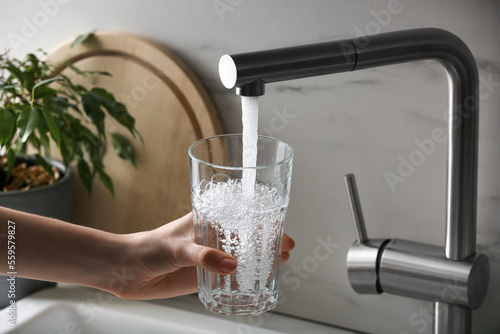 Fotografia Woman filling glass with tap water from faucet in kitchen, closeup