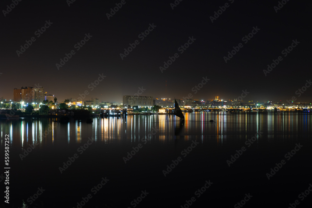 View of the town of Aswan in Egypt, with a Felucca in the middle of the Nile River, and the lights reflected in the water, long exposure photo.