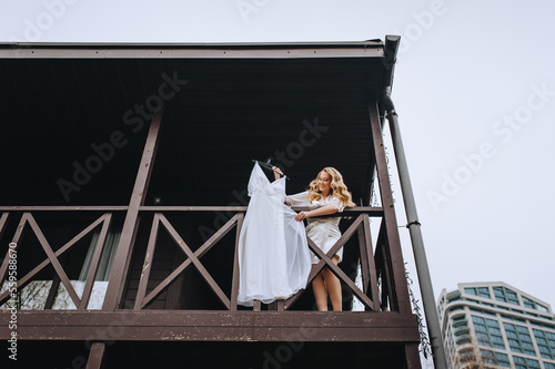 A beautiful smiling curly blonde bride in lingerie, a bathrobe holds a white dress hanging on a hanger, standing on the second floor of a large wooden house, cottage. wedding photography.