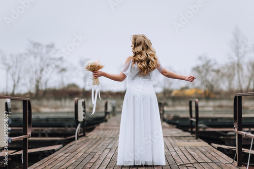 A beautiful curly blonde bride in a white dress stands on a wooden pier in the open air with her back, imitating a bird with her hands. Wedding photography, portrait, autumn.