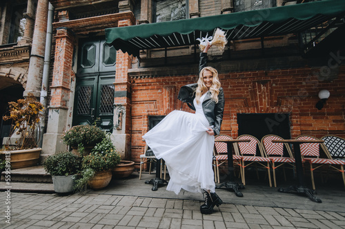 A beautiful curly smiling blonde bride, biker, lover of musical rock in a black leather jacket, white dress with a bouquet of reeds stands, dances in the city. Wedding photography, portrait.