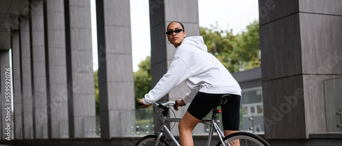 Stylish african american woman riding bike on street, banner view