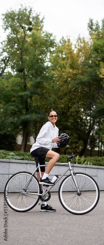 Vertical banner of cheerful african american woman holding helmet near bike on street