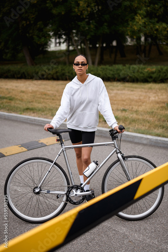 Stylish african american woman standing near bicycle and blurred barrier on road 