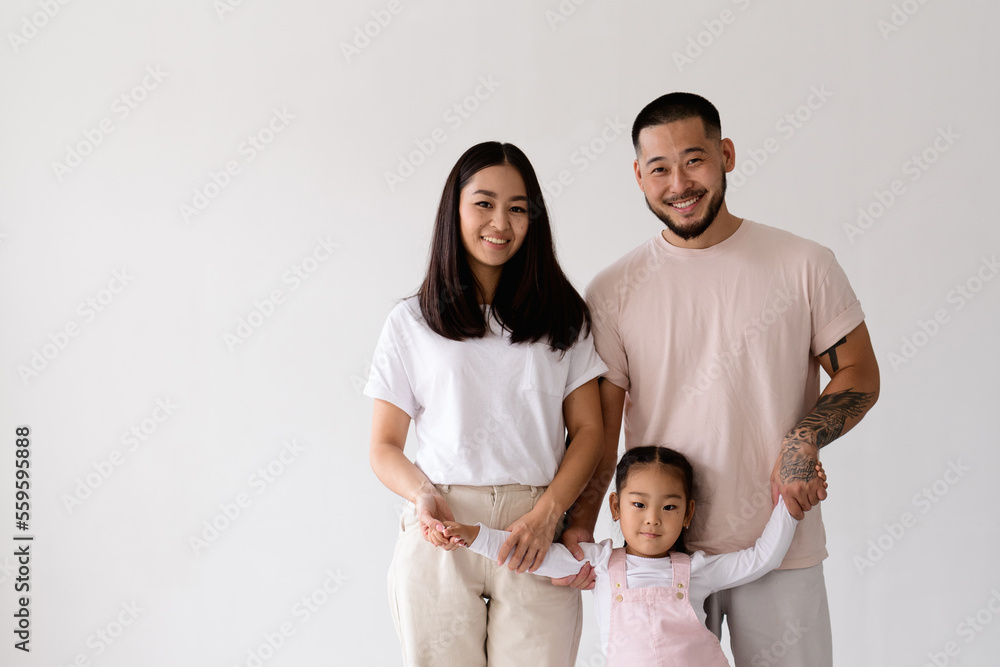 Smiling young asian parents holding hands of toddler daughter and looking at camera isolated on grey 