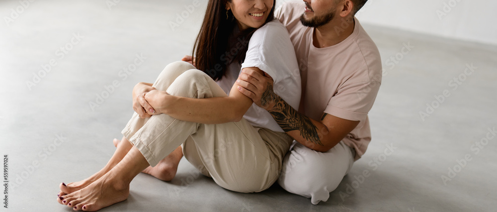 Banner image of smiling tattooed man hugging brunette and barefoot wife while sitting on grey background