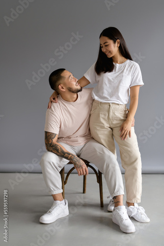 Smiling tattooed man hugging asian wife while sitting on chair on grey background 