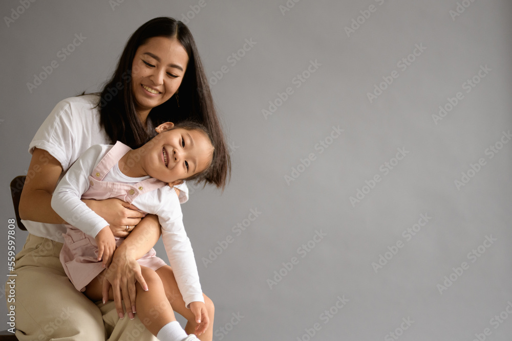 Smiling asian mom hugging toddler daughter isolated on grey background Smiling asian mom hugging toddler daughter isolated on grey background