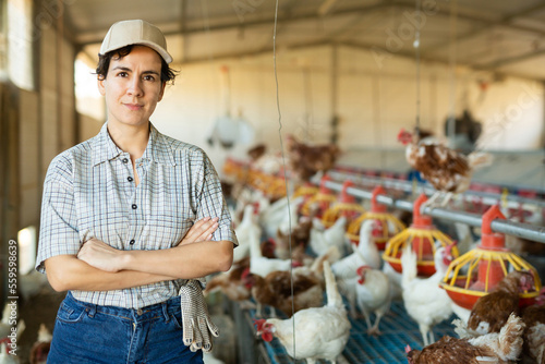 Confident young hispanic woman poultry farm owner standing with arms crossed in henhouse near cages and hanging feeders for laying hens photo