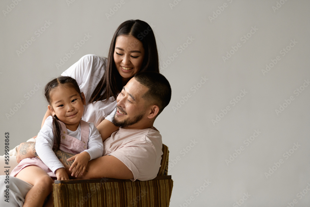 Happy asian parents looking at toddler daughter while sitting on armchair isolated on grey background 