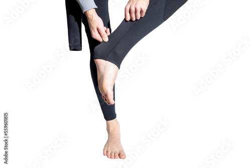 A man putting on a black wetsuit pulling up the legs isolated on a white background. Adjusting a wetsuit balancing on one leg wearing a gray sweatshirt. 