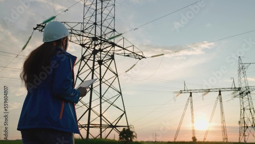 Woman Electricity supply company employee works outdoors, services high voltage electric lines at sunset. Engineer woman power engineer in white helmet checks power line using computer tablet