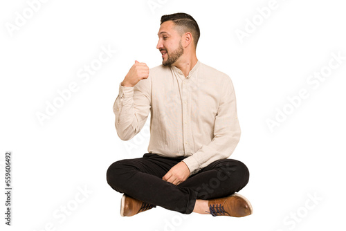 Adult latin man sitting on the floor cut out isolated points with thumb finger away, laughing and carefree.