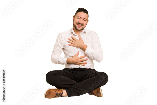 Adult latin man sitting on the floor cut out isolated laughs happily and has fun keeping hands on stomach.