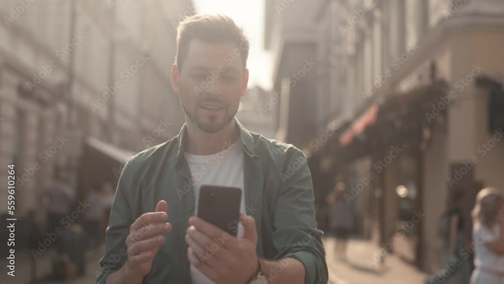 Portrait happy affable attractive caucasian man video calling people using smartphone online smiling. Beautiful young adult male talking communicating standing in picturesque city center on sunny day. Portrait happy affable attractive caucasian man video calling people using smartphone online smiling. Beautiful young adult male talking communicating standing in picturesque city center on sunny day.