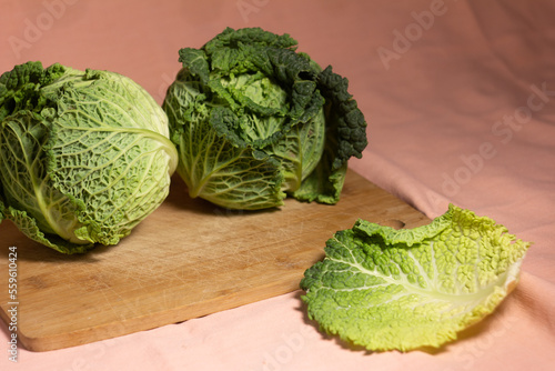 Two heads of savoy cabbage on a wooden bamboo cutting board on a pink background with one leaf lying apart