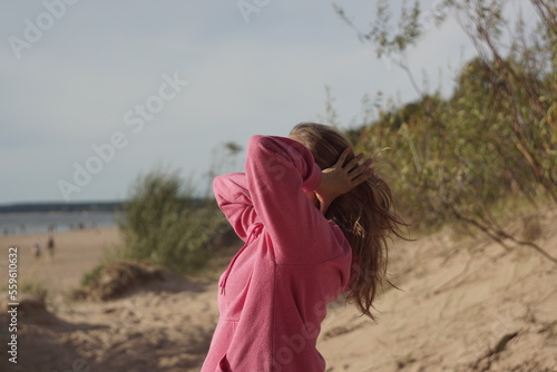 Young woman alone at a beach looking at water while fixing her hair. She is enjoying being outside in nature. Woman is wearing pink sweatshirt. Her face is not visible.
