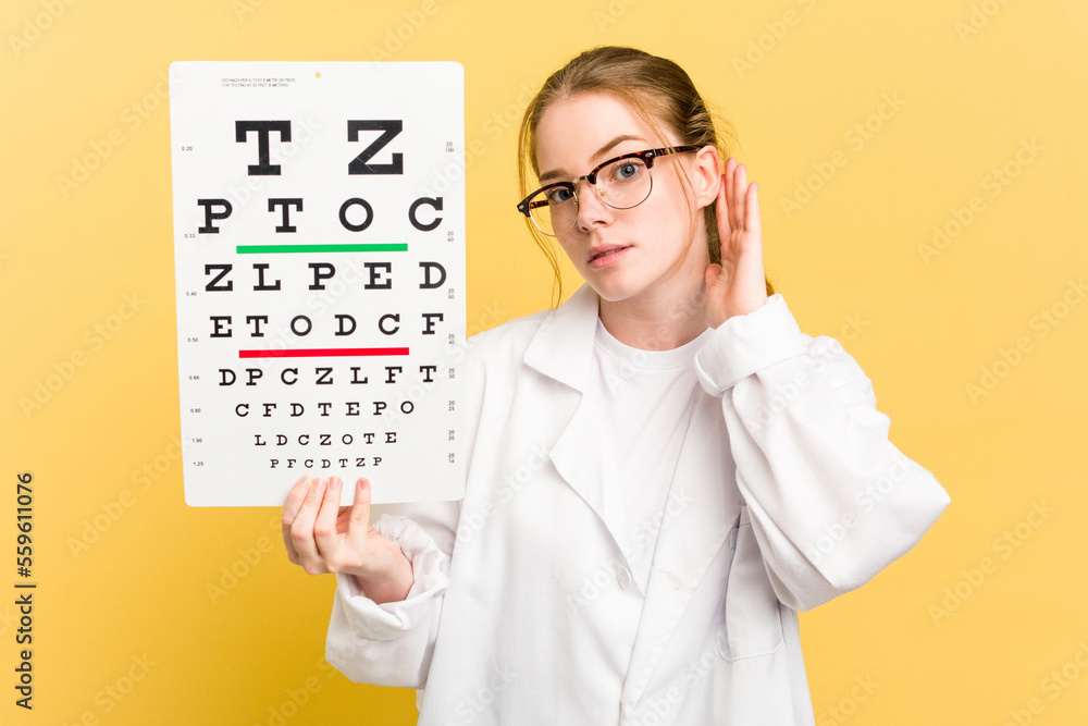 Young caucasian oculist woman holding a eye chart paper isolated on yellow background trying to listening a gossip. Young caucasian oculist woman holding a eye chart paper isolated on yellow background trying to listening a gossip.