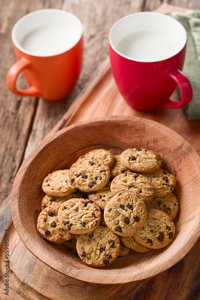 Chocolate chip cookies in wooden bowl with two cups of milk in the back, photographed on wood (Selective Focus, Focus one third into the bowl) Chocolate chip cookies in wooden bowl with two cups of milk in the back, photographed on wood (Selective Focus, Focus one third into the bowl)