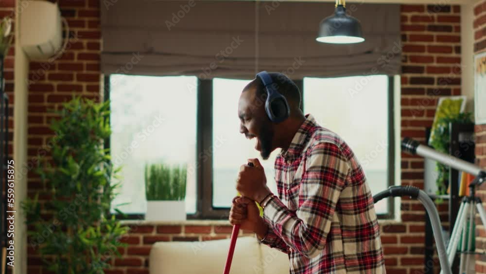 Cheerful young adult mopping floors with washing solution, dancing and listening to music on headset. Modern happy man having fun cleaning living room, using mop to wash and sweep dirt. Cheerful young adult mopping floors with washing solution, dancing and listening to music on headset. Modern happy man having fun cleaning living room, using mop to wash and sweep dirt.