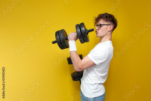 young thin, not athletic, nerd guy lifts heavy dumbbells and shows strength on a yellow background