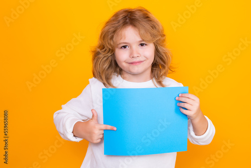 Kid boy with sheet of paper, isolated on yellow background. Portrait of a kid holding a blank placard, poster. Empty board for templates banner, copy spase, mock up.