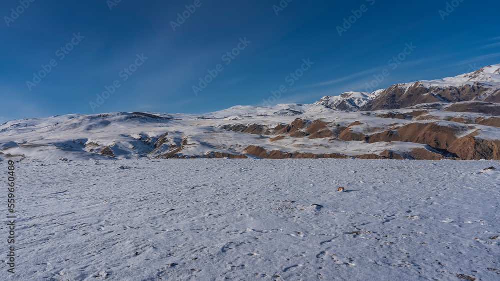 A mountain range with red-brown slopes against a blue sky. Footprints are visible in the snowy valley. Altai Mars. Kyzyl Chin
