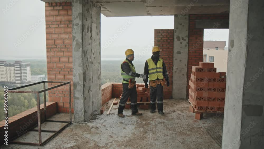 Long shot of ethnically diverse men wearing uniform working at multistory building under construction discussing materials and worktools Long shot of ethnically diverse men wearing uniform working at multistory building under construction discussing materials and worktools