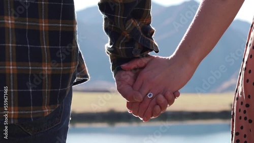 Couple in love holding hands after getting engaged with engagement ring on finger looking out onto beautiful lake view