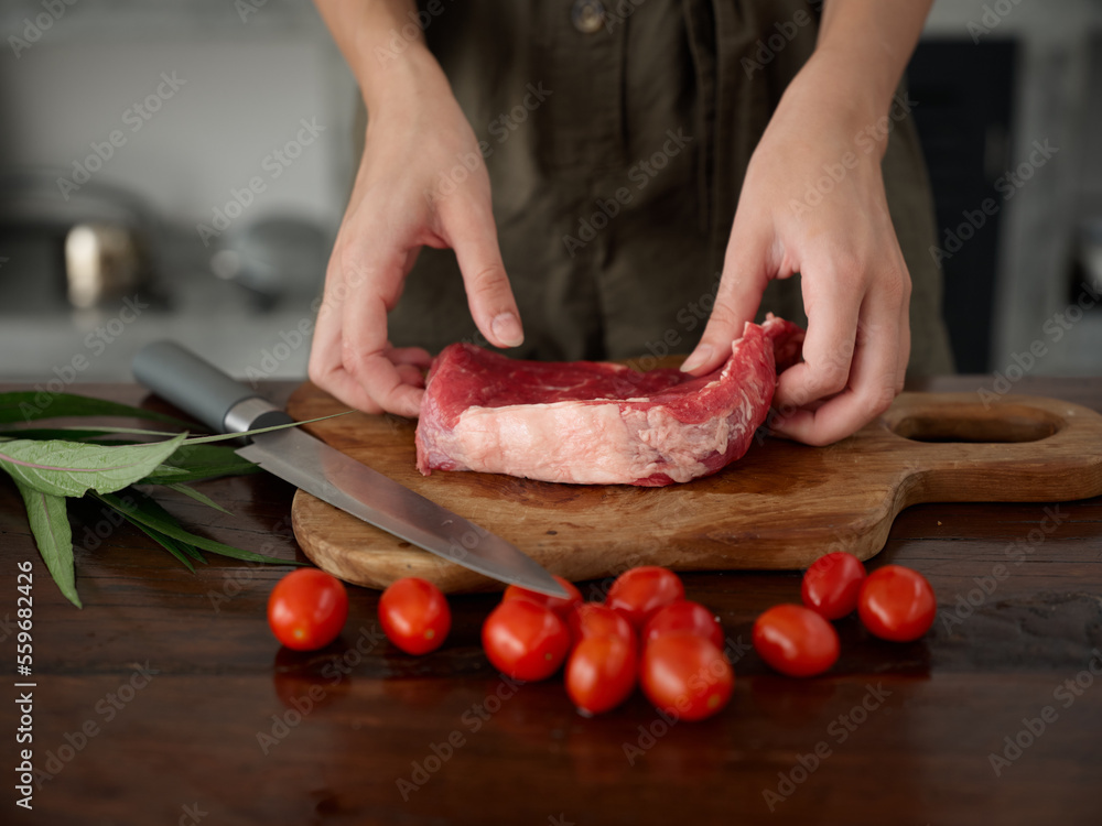 Woman with knife in hand cutting fresh steak meat for roasting in kitchen with salt pepper and other spices on table, red cherry tomatoes and herbs, preparing dinner. Woman with knife in hand cutting fresh steak meat for roasting in kitchen with salt pepper and other spices on table, red cherry tomatoes and herbs, preparing dinner.