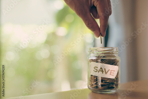 Hand putting Coins in glass jar with SAVE word written text label for Education, Necessity, Play, Long Term, Give and Financial Freedom concept.