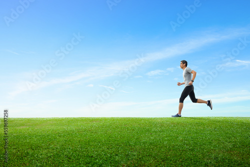 Side view of asian man jogging on spring grass field with clear blue sky background.