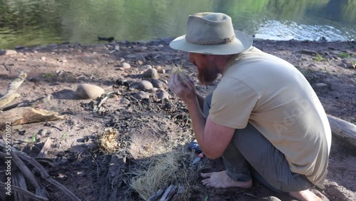 A bushman in an akubra hat creates fire by blowing an embar in grass in the Australian bush.