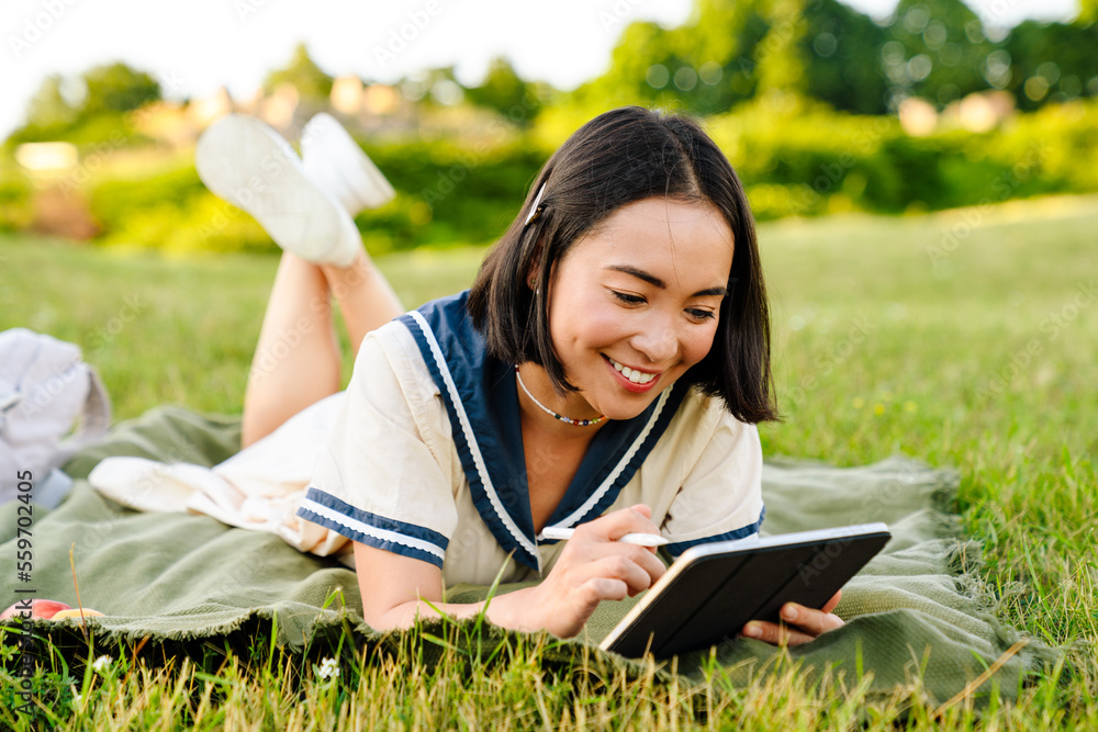 Smiling asian woman drawing on digital tablet with stylus while lying on blanket in park Smiling asian woman drawing on digital tablet with stylus while lying on blanket in park