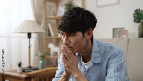 depressed asian young man sitting on the living room sofa in silence and showing no expression on face while staying at home alone during self isolation.