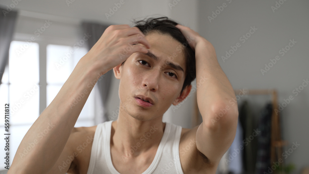 cheerful asian Korean man looking at the camera as mirror is combing his hair with hand and smiling while going through morning beauty routine in the bedroom at home. cheerful asian Korean man looking at the camera as mirror is combing his hair with hand and smiling while going through morning beauty routine in the bedroom at home.
