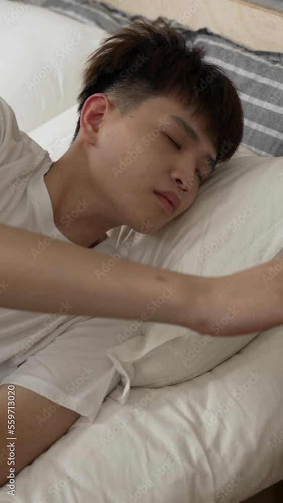 Vertical Video: asian man hitting the snooze button on the alarm clock and keep sleeping on bed in the morning in the bedroom at home. Vertical Video: asian man hitting the snooze button on the alarm clock and keep sleeping on bed in the morning in the bedroom at home.