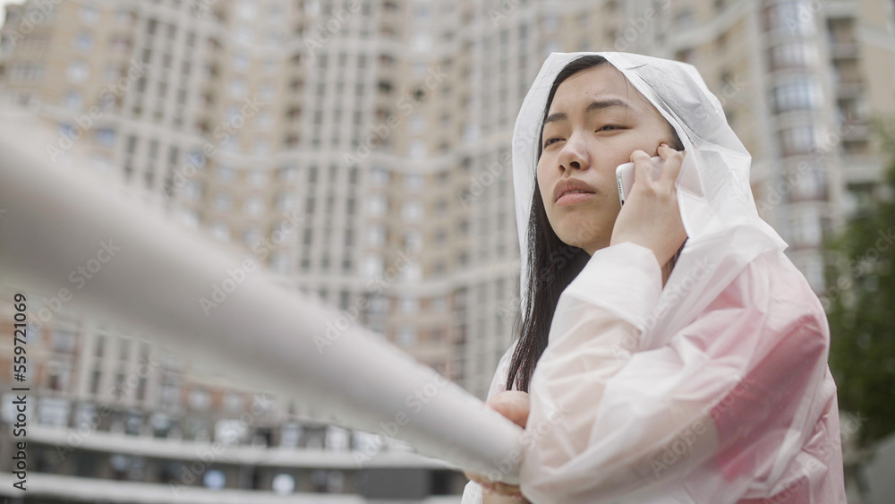 Fototapeta premium Upset asian woman in raincoat talking on phone in rainy city, calling taxi