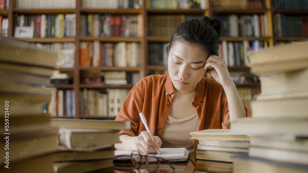 Tired asian female student working on essay at university library, lots of books Tired asian female student working on essay at university library, lots of books