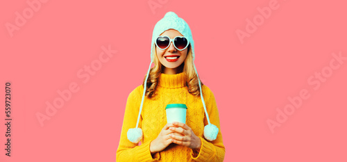 Winter portrait of happy smiling young woman with coffee cup wearing yellow knitted sweater, white hat with pom pom, heart shaped sunglasses on pink background